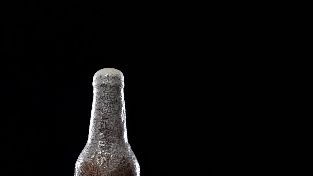 Liquor splashes out when cold bottle of beer opening, foam bubbles rising and overflowing. Beer foam flows down a misted bottle on dark background