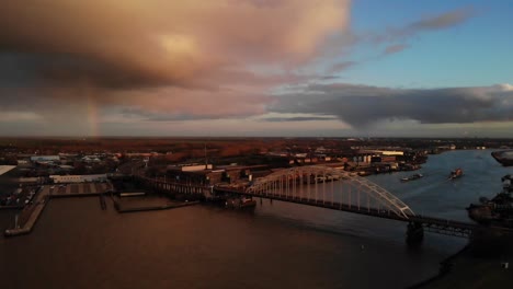 bird's eye view of arch bridge over noord river with barge and freight ship sailing during sunset with rainbow in hendrik-ido-ambacht, netherlands. - aerial, static