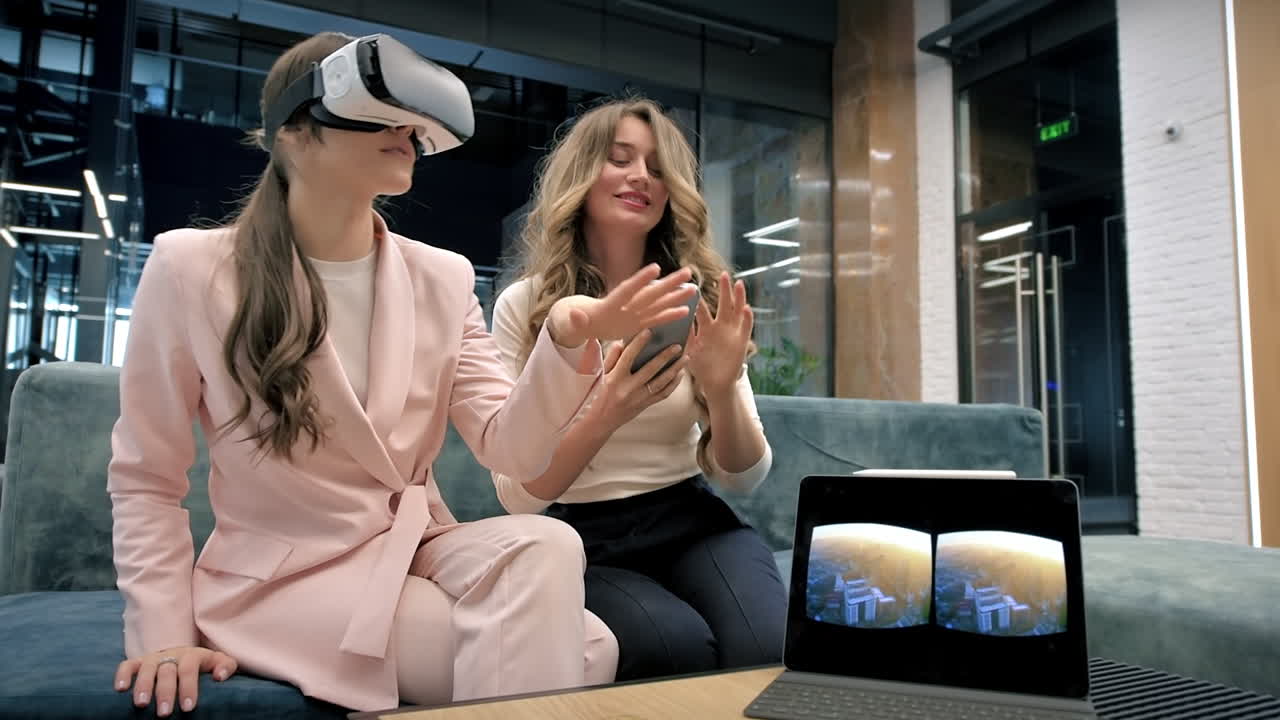A blonde caucasian woman is using VR glasses in an office while another woman is using smartphone, both listening music. Tablet on the table showing image visible in the headset