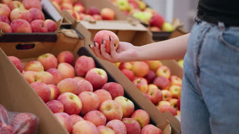 close-up of woman selecting fresh red apple from a pile of apples in a cardboard box at a grocery store, showcasing hand with black nails reaching for fruit