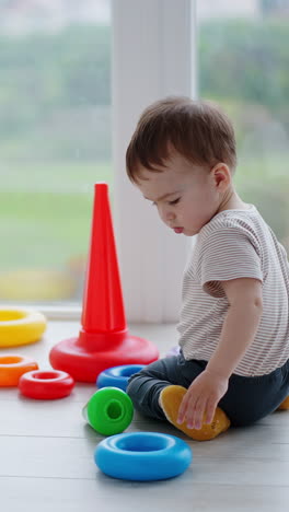 caucasian baby boy sits on the light balcony near his toys. kid lazily plays with a pyramid. vertical video