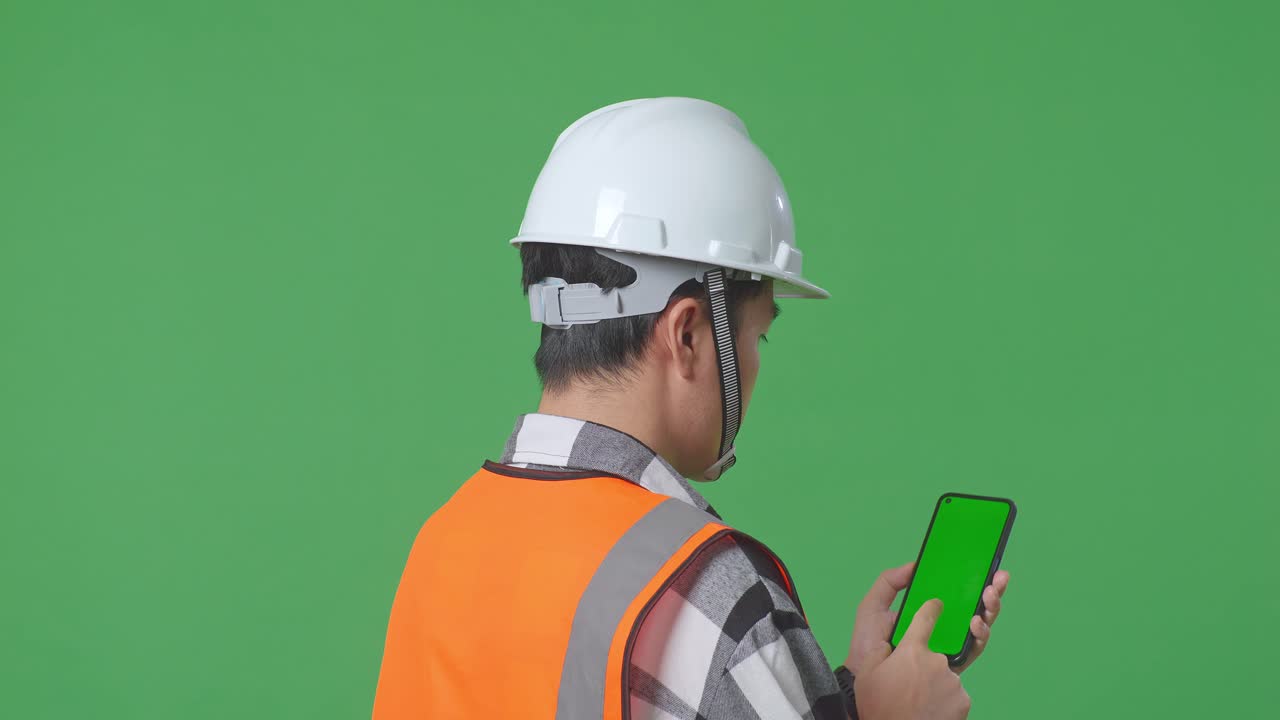 Close Up Back View Of Asian Male Engineer With Safety Helmet Working On A Green Screen Smartphone And Looking Around While Standing In The Green Screen Background Studio