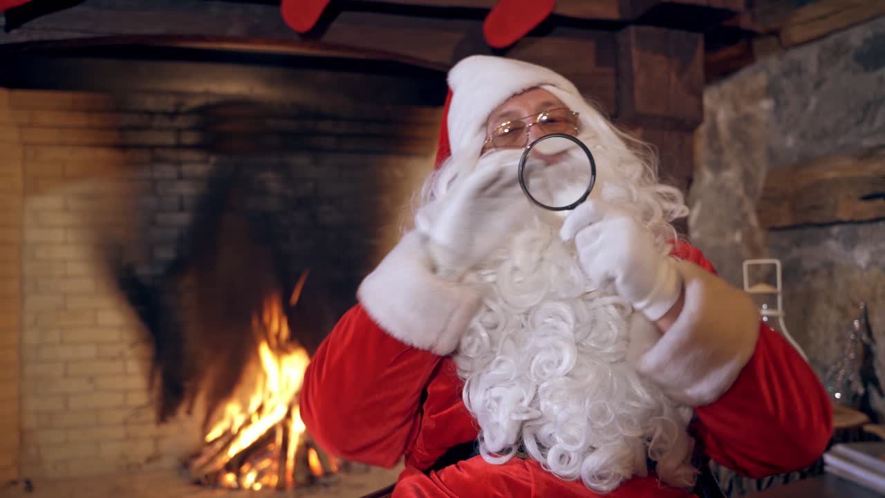 Santa Claus with magnifying glass. Real Santa in red costume with white beard holding magnifying glass on the fireplace background. Christmas spirit.