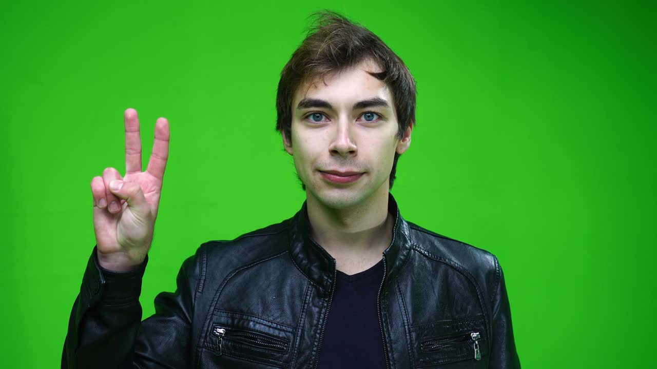 가죽 재을 입은 백인 남자가 녹색 배경에 승리 또는 평화 표지판을 보여줍니다.