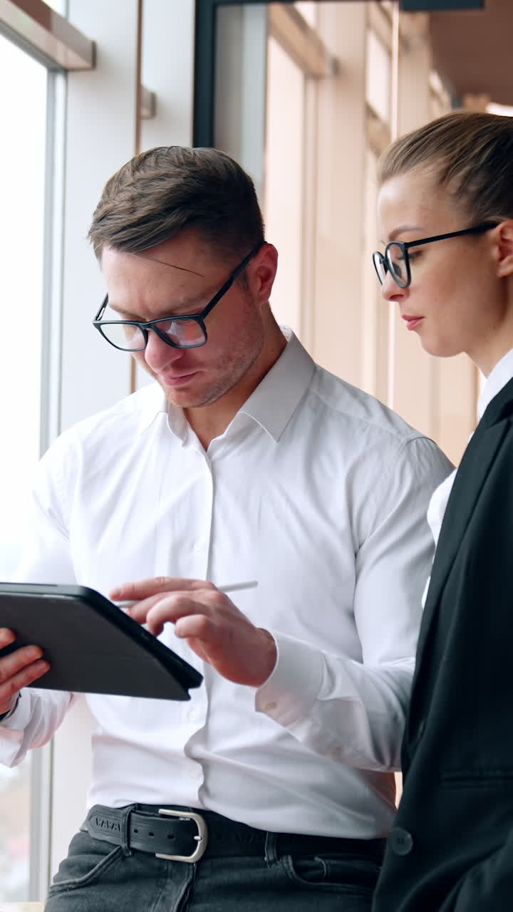 Focused colleagues wearing glasses look at I-pad in male hands. Both co-workers look at window and smile. Vertical video