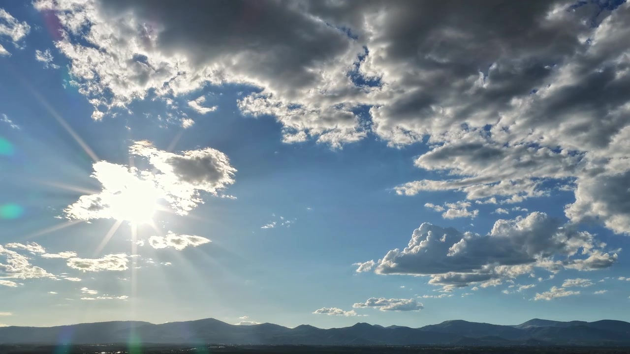 Un timelapse 4K realizzato con drone che cattura il sole splendente con brillanti bagliori dell'obiettivo e raggi di luce che si spostano accanto alle nuvole sopra il sereno paesaggio della catena montuosa di Jemez