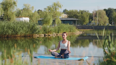 caucasian young woman sitting on mat in lotus position relaxing near pond in park on summer day