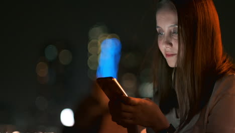 por la noche, una joven se sienta en la hierba de una gran ciudad y mira a la pantalla de su teléfono inteligente, escribe un mensaje y mantiene su blog personal.