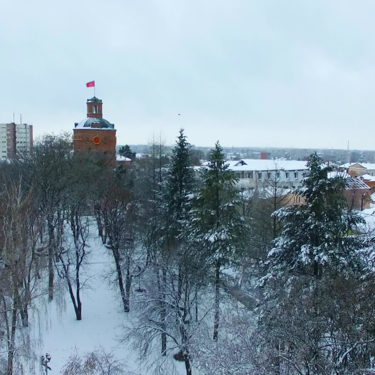 Park in winter season with old tower with clock. Drone rising over the snowy city panorama at daytime. Top view