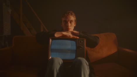 portrait of young man sitting on couch, holding retro tv in dimly lit room