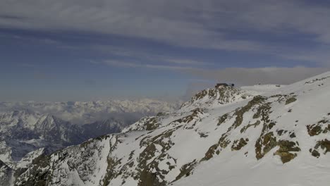 Drone-flying-close-to-snowy-mountain,-heading-towards-Rifugio-Quintino-Sella-al-Felik,-with-backdrop-of-the-Alps-and-Mont-Blanc-Massif-in-the-distance