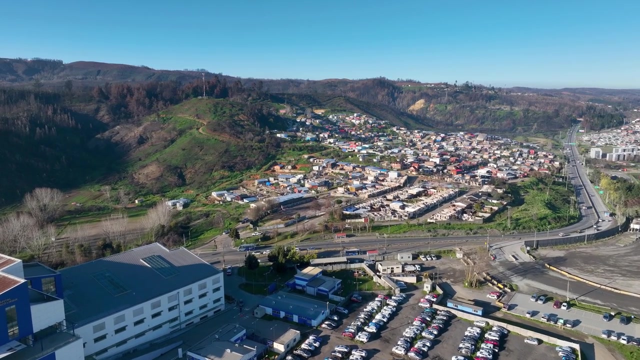 Volo in avanzamento su una densa città collinare con tetti colorati e strade tortuose che attraversano colline bruciate ed edifici industriali sotto un cielo azzurro limpido. Lirquen, Penco, Cile