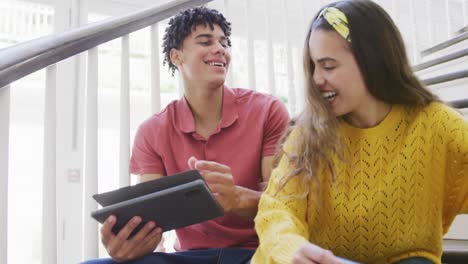 una pareja biracial feliz pasando tiempo en casa usando una tableta, abrazándose y sentándose en las escaleras.