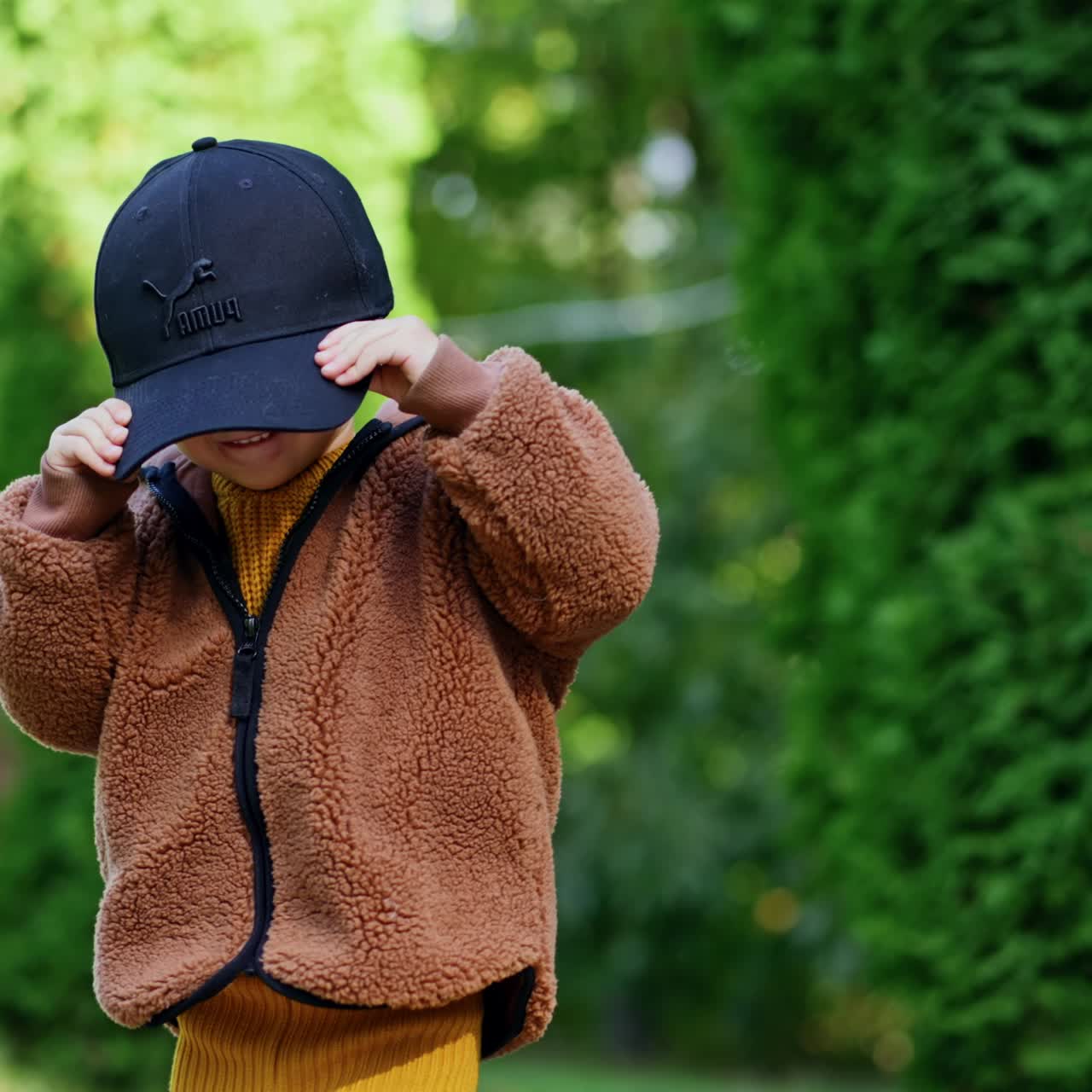 Funny little kid playing with a black cap too big for him. Cute baby boy in the green park