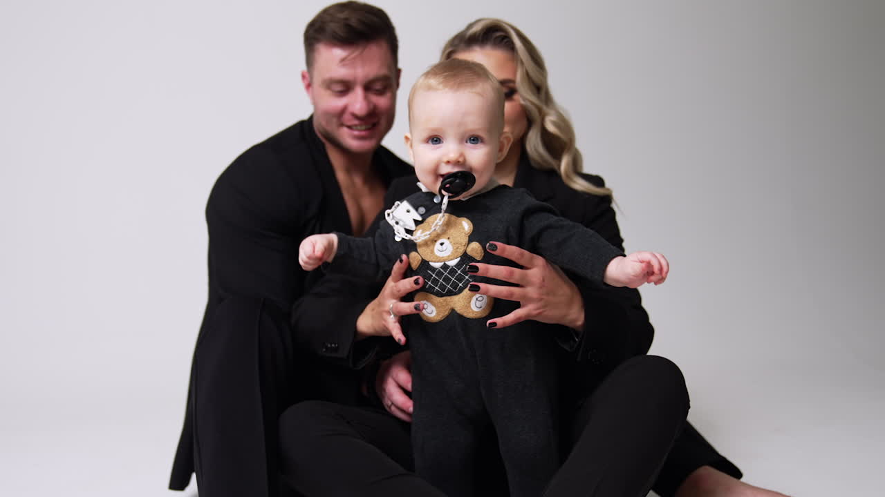 Proud young parents with their lovely infant baby. Cute child with pacifier in mouth tries to stand himself. White backdrop.