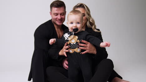 proud young parents with their lovely infant baby. cute child with pacifier in mouth tries to stand himself. white backdrop.