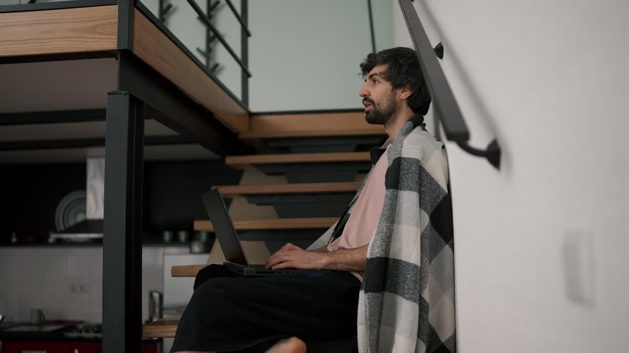 joven barbudo caucásico trabajador independiente usar computadora portátil, sentado en escaleras de madera en casa en cuadros