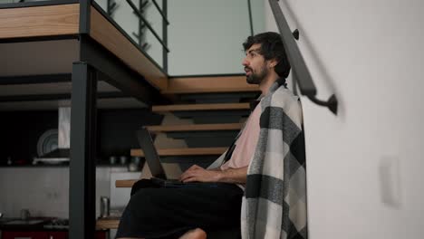 young bearded caucasian freelance worker use laptop, sitting on wooden stairs at home in plaid