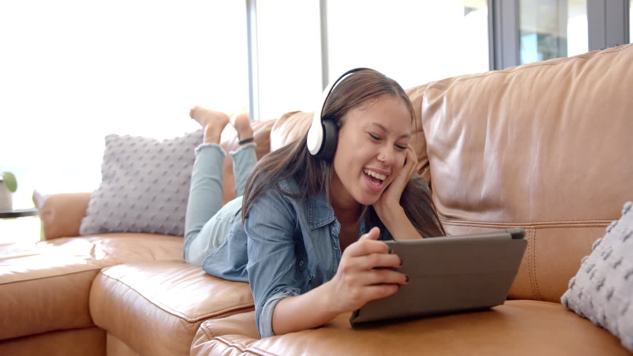Listening to music with headphones, girl laughing while using tablet on couch