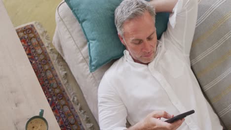 Happy-caucasian-man-lying-on-sofa-in-living-room,-using-smartphone