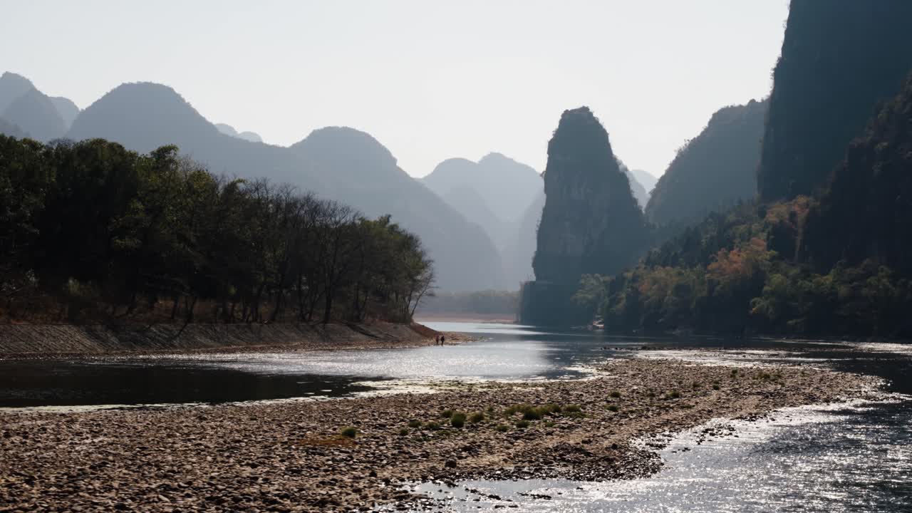 Sunlit water sparkles over rocky banks as hazy karst mountains rise along the Li River