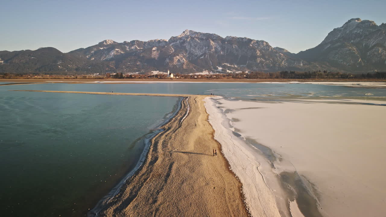 La telecamera avanza lungo una stretta lingua di sabbia dove neve e ghiaccio incontrano acque blu intenso, con le montagne bavaresi e un villaggio invernale in lontananza