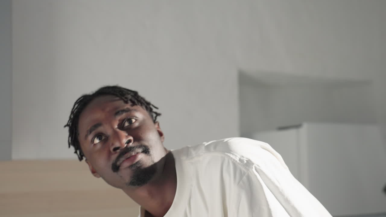 Man looking up thoughtfully while standing in modern kitchen, wearing casual white shirt and black shorts. Soft lighting and neutral background create a calm and reflective atmosphere