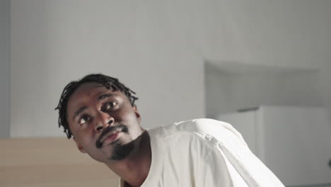 man looking up thoughtfully while standing in modern kitchen, wearing casual white shirt and black shorts. soft lighting and neutral background create a calm and reflective atmosphere