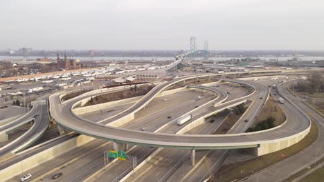 Winding-road-and-Ambassador-bridge-connecting-USA-and-Canada-with-traffic-of-semi-trucks,-aerial-view