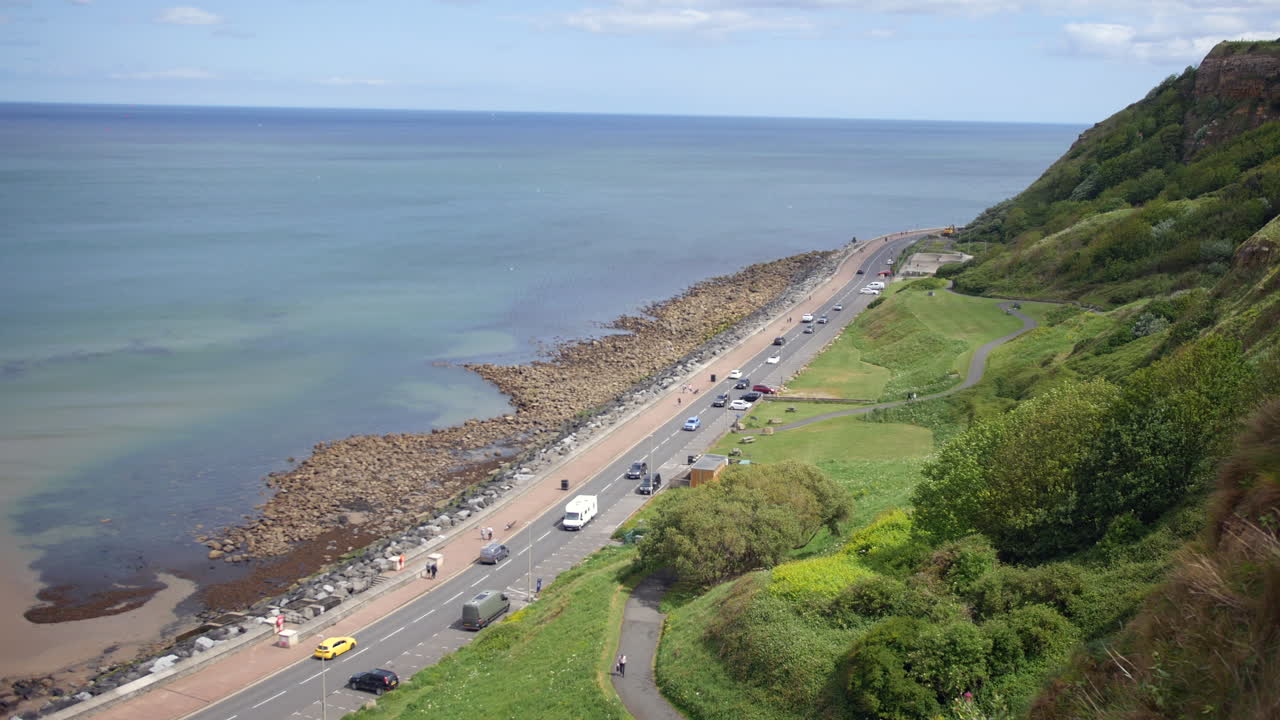 A coastal road curves along the shoreline in Scarborough, North Yorkshire, England, with cars driving past rocky beaches and grassy cliffs under a clear sky