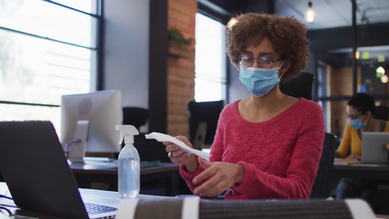 African american woman wearing face mask cleaning her smartphone using disinfectant spray at modern