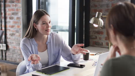 Young-Caucasian-woman-in-a-blue-shirt-converses-with-an-Asian-woman-in-a-business-office-setting