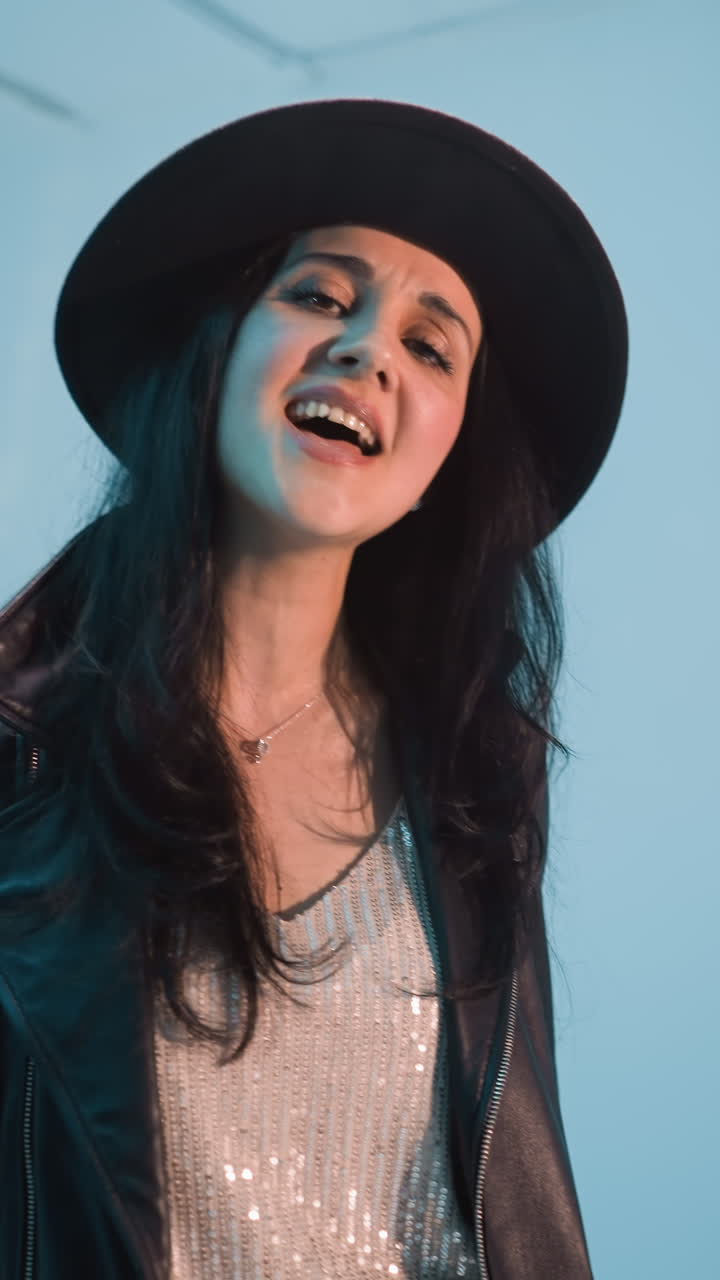 una mujer con una chaqueta de cuero negra, pantalones y sombrero canta con pasión, moviendo su cuerpo y manos expresivamente contra un fondo azul. ella actúa sin un micrófono