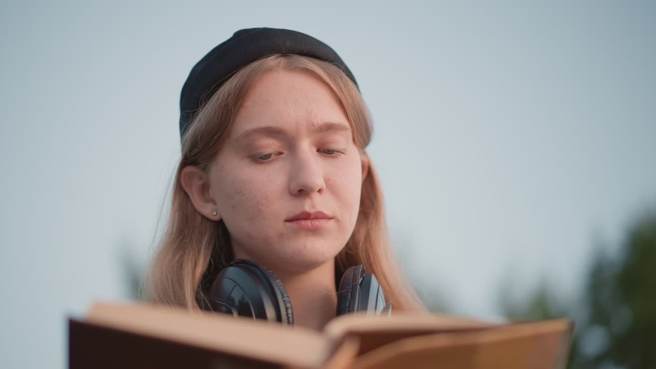 primo piano crepuscolare studente bianco con cuffie che legge, sessione di studio intensa tra pagine aperte e libro con copertina rigida, espressione attenta, note a margine e segnalibro visibili, campus esterno tranquillo