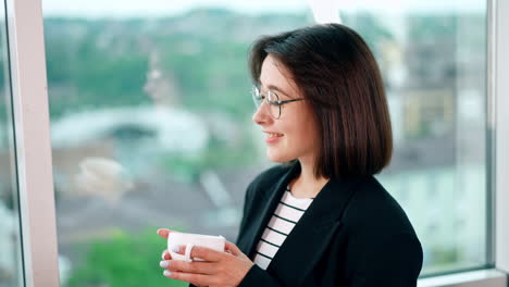 beautiful smiling lady wearing glasses holds a white cup. woman turn to look at the window taking a sip of coffee.