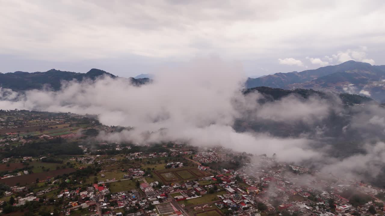 Thick fluffy clouds hover over a village with red roofs, nestled near the mountains.