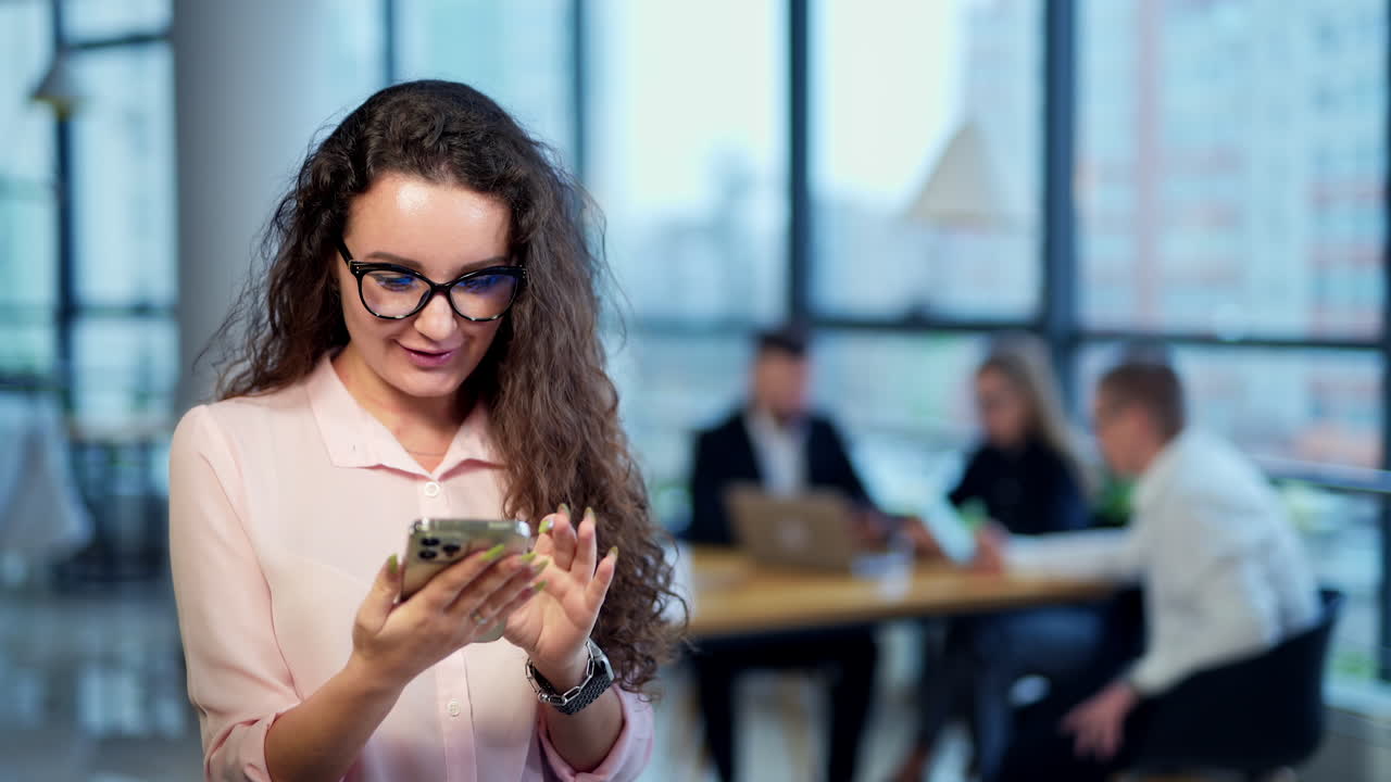 Beautiful lady holding a phone and searching something there in a very good mood. Black-haired woman messaging to someone. People at the table in blur at the backdrop.