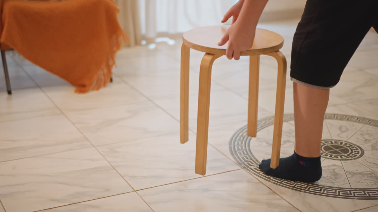 Small wooden stool on tiled floor, person steps forward and picks up stool, patterned medallion tile underfoot, orange throw on sofa, natural morning light, gentle domestic action in simple interior