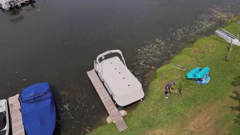 Man-Cutting-Grass-With-Grass-Cutter-On-The-Riverbank-Near-Boat-Dock