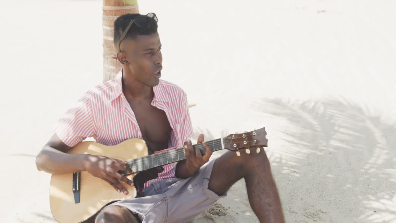 hombre afroamericano sentado bajo un árbol, tocando la guitarra y cantando en una playa soleada, en cámara lenta.