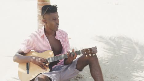 African-american-man-sitting-under-a-tree,-playing-guitar-and-singing-on-sunny-beach,-slow-motion