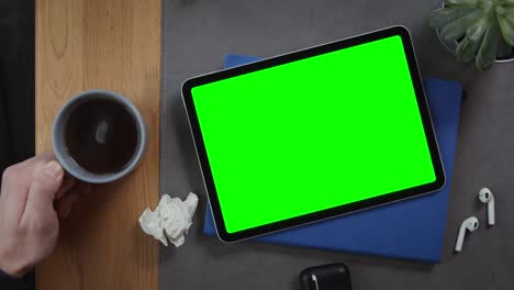 a man puts cup of tea on the desktop next to a tablet with a green screen