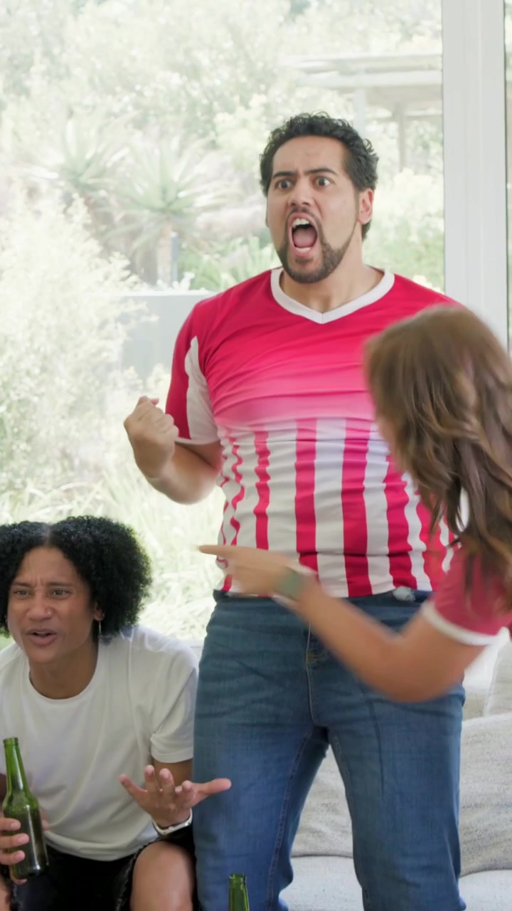 Vertical video: Reacting to game on TV man in red jersey standing celebrating with beer at home