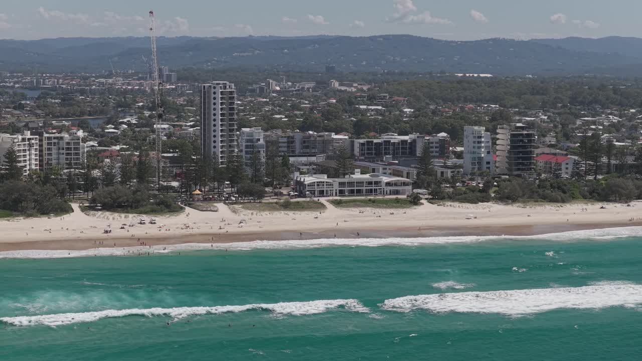 Menschen auf dem weißen Sand des North Burleigh Beach mit Küstenapartments und Bergen im Hinterland. Weite Luftaufnahme.