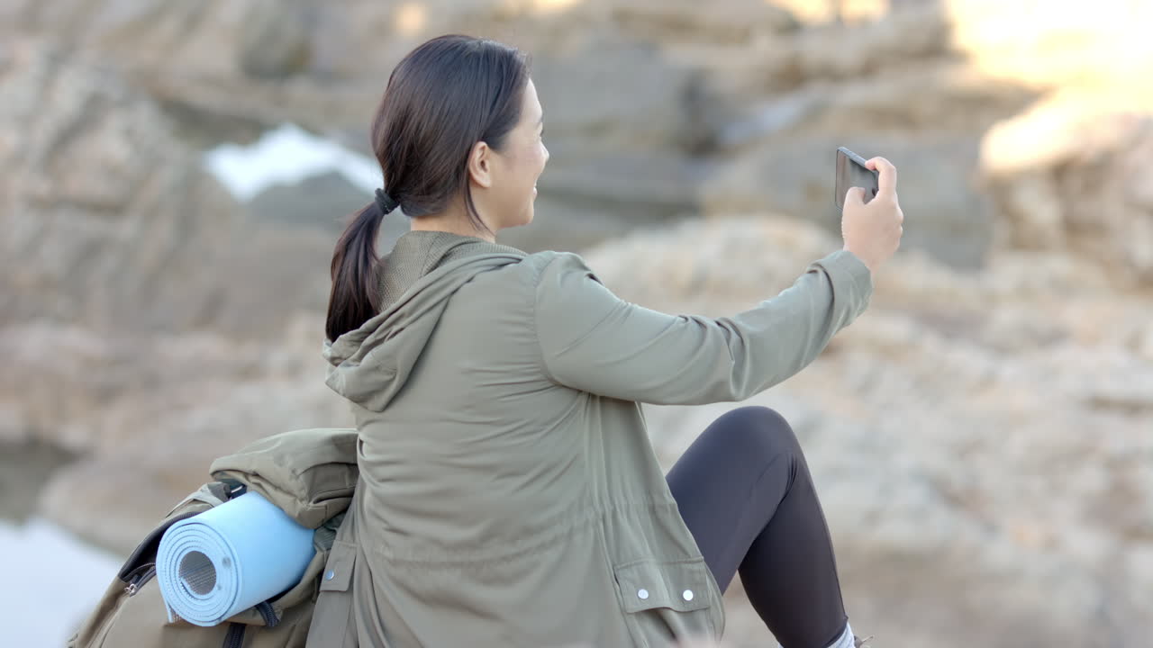 Taking selfie, woman sitting on rock with yoga mat during mountain hike