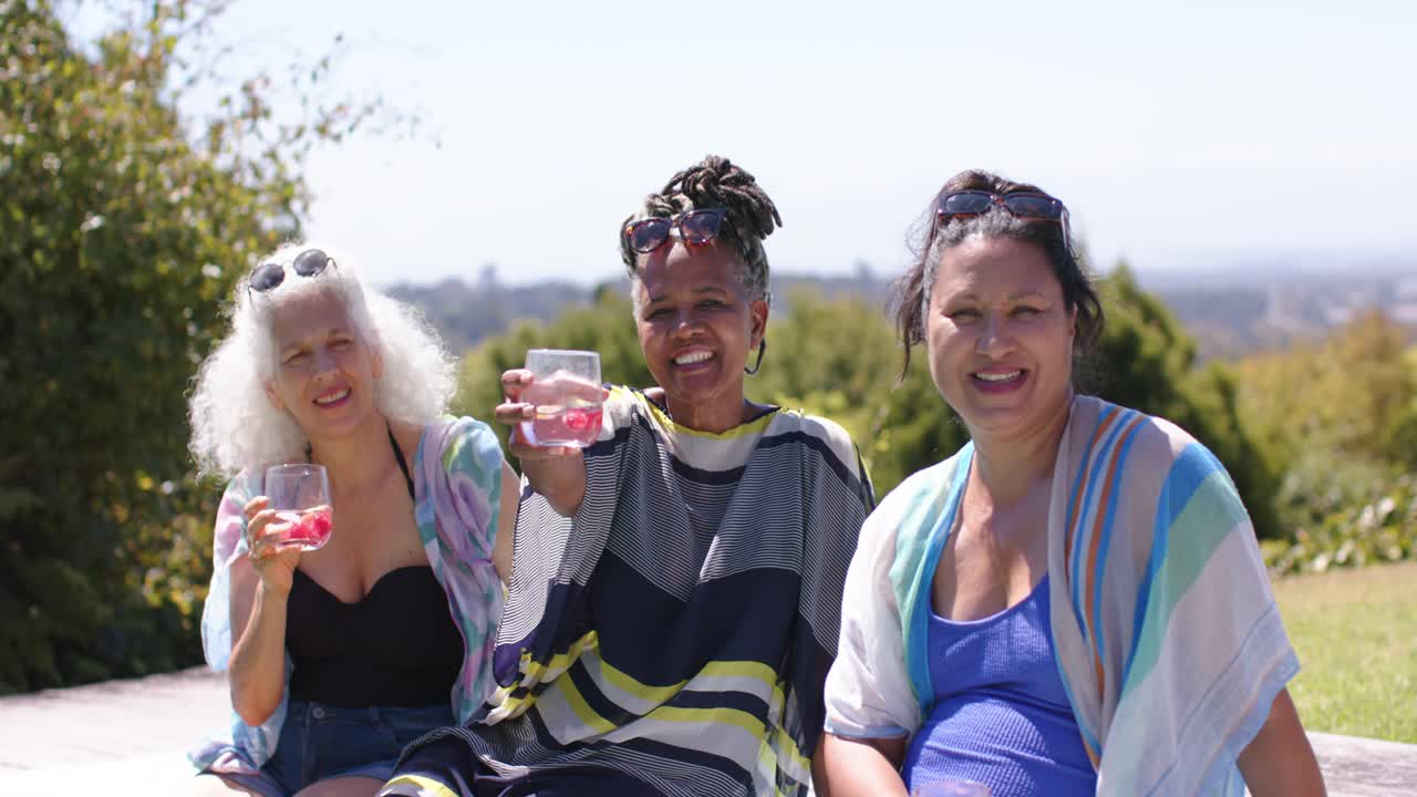 Trio femminile di diverse etnie in costume da bagno che brindano con bevande rosa, mentre la telecamera si allontana a bordo piscina