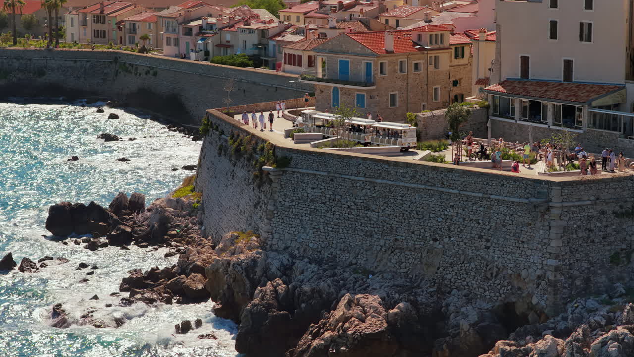 Aerial drone view of Antibes stone ramparts and seaside promenade with visitors walking along the walls, overlooking the rocky Mediterranean coast