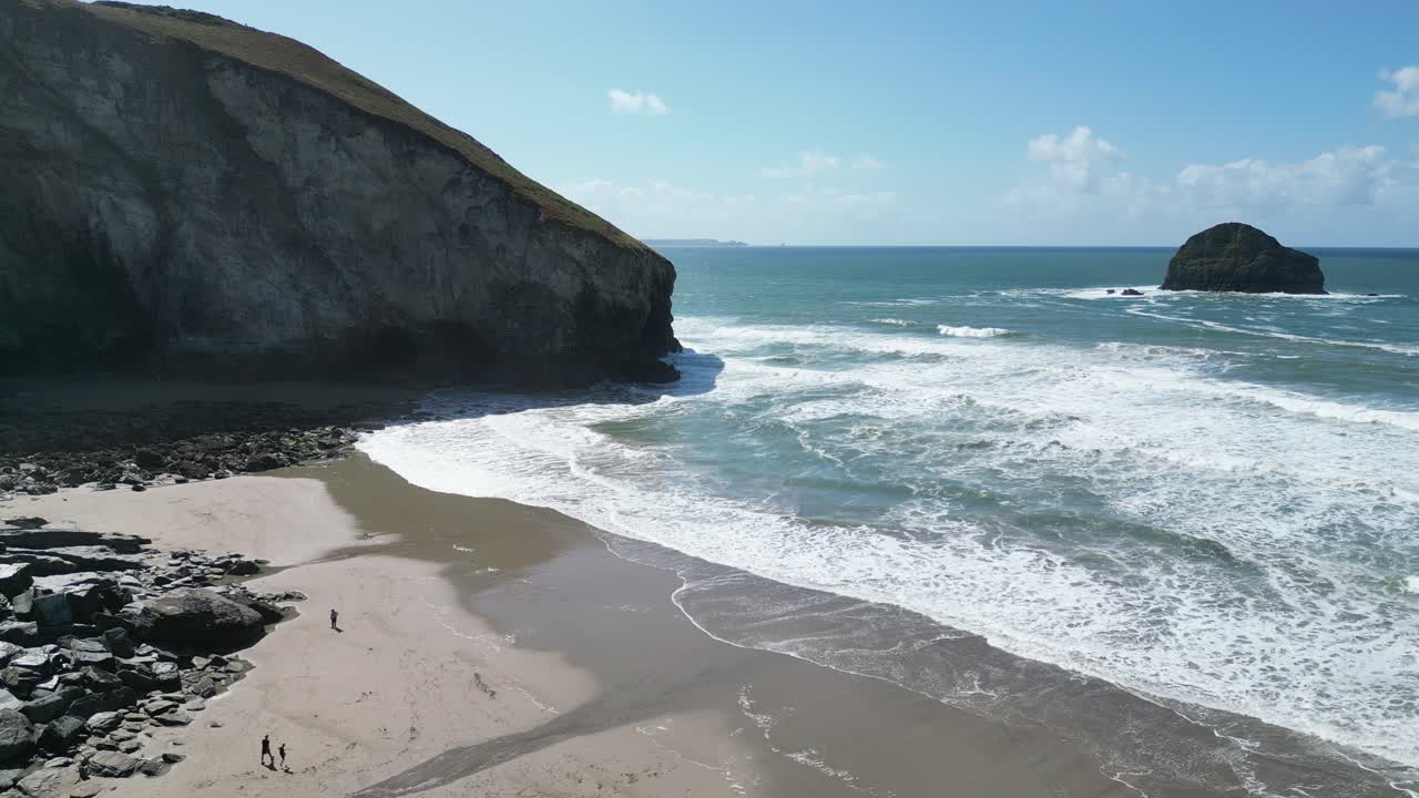 Stunning aerial view shows Trebarwith Strand coastline in Cornwall with dramatic cliffs, rocky formations, sandy beach, and powerful ocean waves under clear blue sky