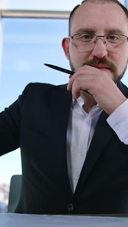 portrait of positive businessman. successful entrepreneur sitting at table and looking at camera. bearded boss in glasses and elegant suit in office. vertical video