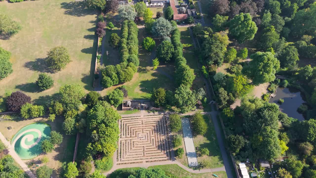 Drone flyover starting at the maze and Venusberg in Cedar’s Park, Cheshunt, England, passing over buildings to the main road, with vibrant summer trees